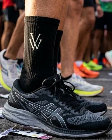 Person wearing black running shoes and socks with a logo at a race start line.